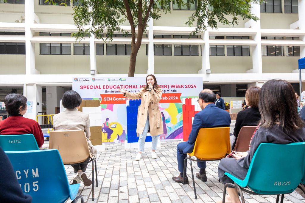 Andrea Hosking, a fourth-year psychology major at The University of Hong Kong, sings in front of an audience in an outdoor setting. Behind her, a banner reads 