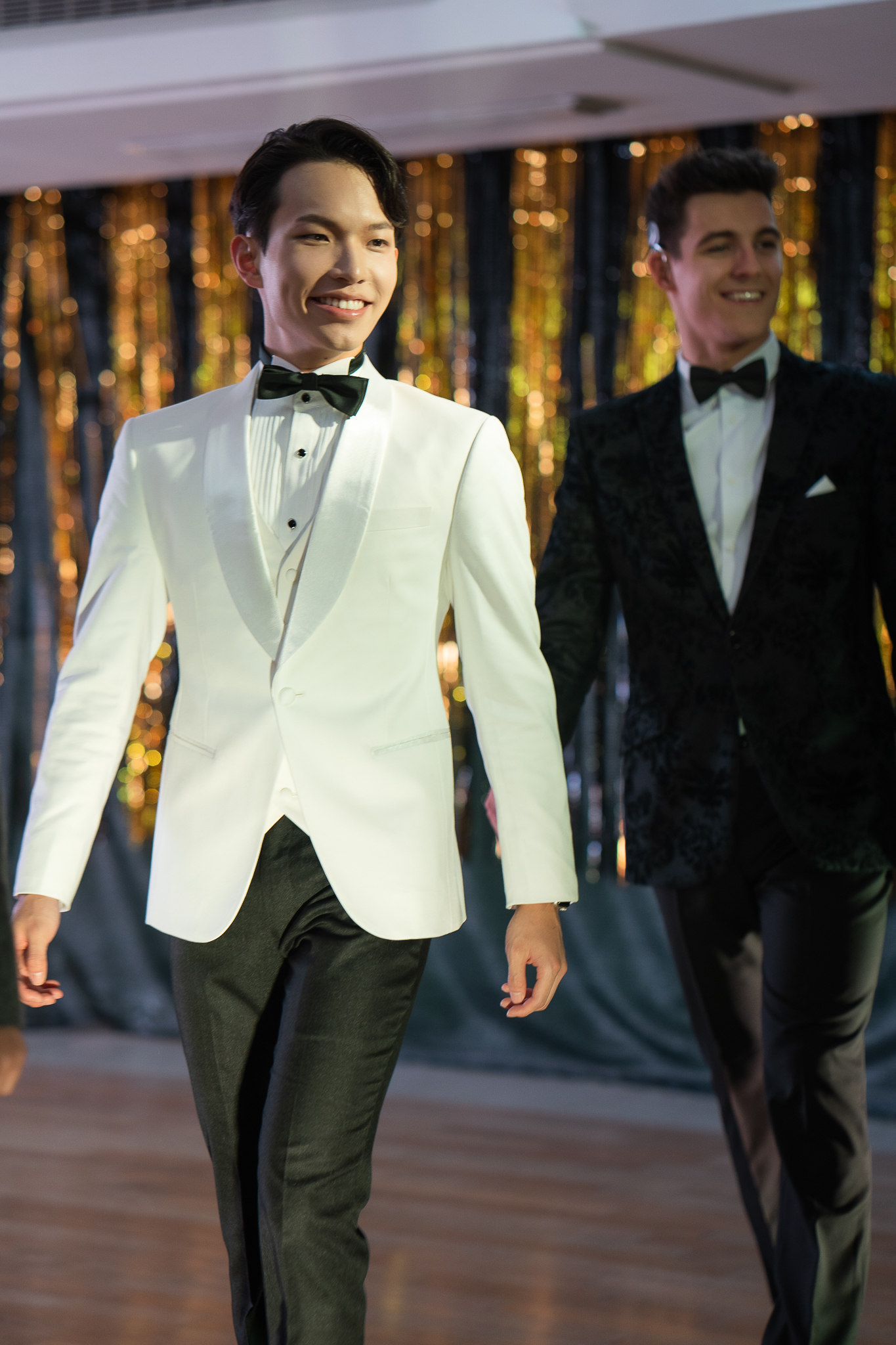 Marco, wearing a white tuxedo jacket, black pants, and a black bow tie, walks on a wooden floor in front of a backdrop with vertical golden and black stripes. Another person in a dark tuxedo jacket with a subtle pattern walks beside him.