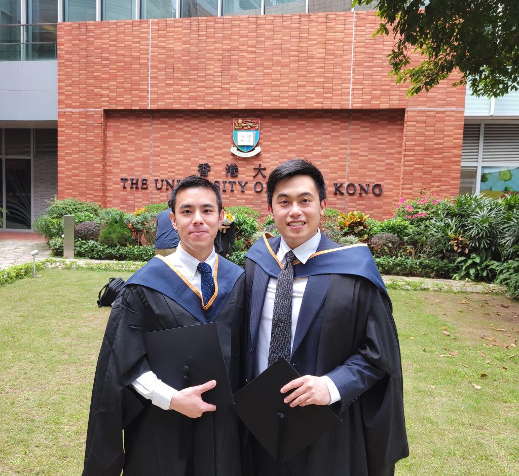 Nathan Fok and his classmate in graduation gowns and caps are standing side by side, holding their diplomas. They are in front of a brick building with the text 'THE UNIVERSITY OF HONG KONG' displayed prominently on it. The background includes greenery and part of the building's facade. 