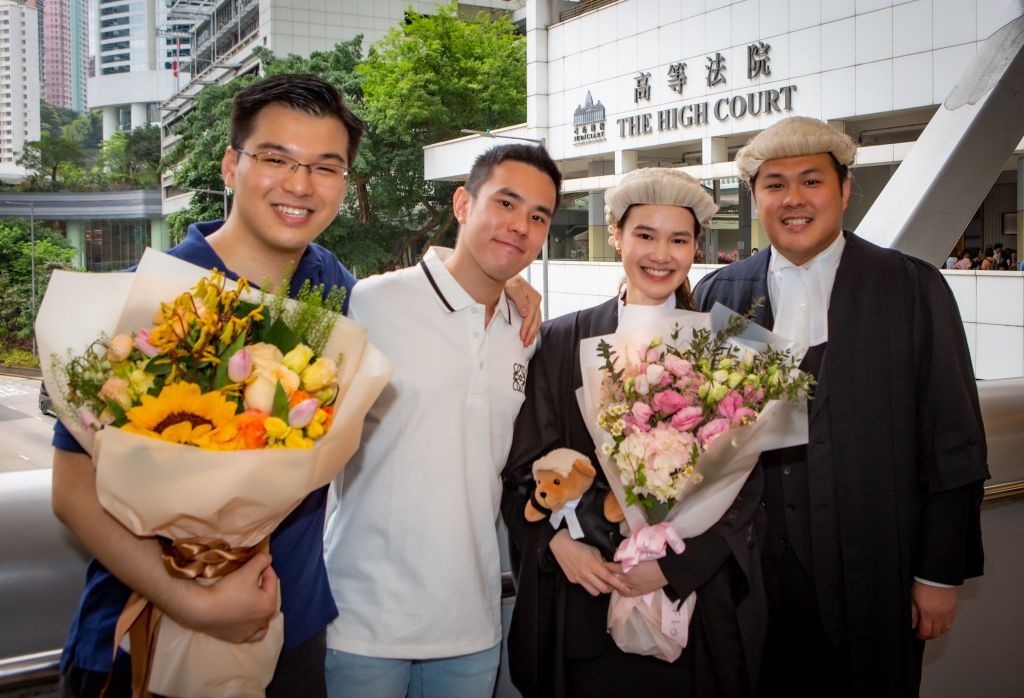A group of four people standing in front of a building with a sign that reads 'THE HIGH COURT.' The person second from the left, Nathan, is wearing a white T-shirt. Two people on the left are holding bouquets of flowers. The person on the far right is dressed in formal attire, possibly a judicial robe.